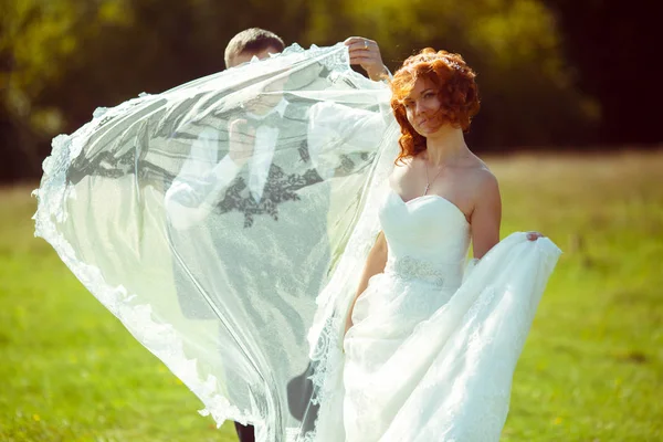 Vőlegény menyasszony fátyol mögött bújik meg míg ő pózol zöld mezőn — Stock Fotó