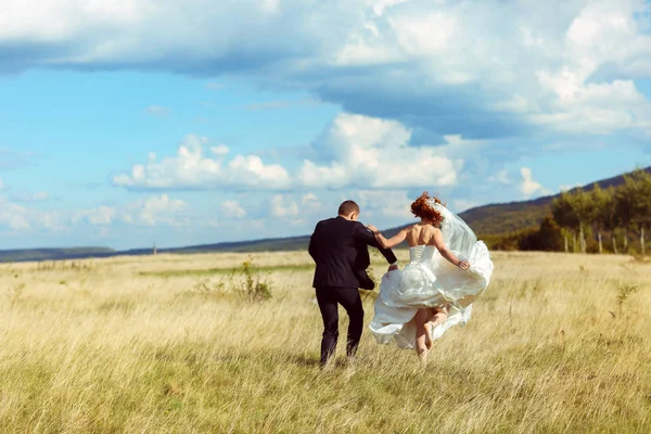 Novia salta caminando detrás de un novio acroos un campo de oro — Foto de Stock