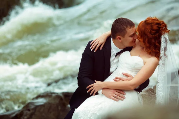 Groom beija uma noiva sentada atrás de um rio de montanha — Fotografia de Stock