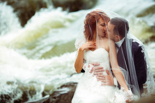 Bride kisses groom hidden under her veil, sitting on the shore of mountain river