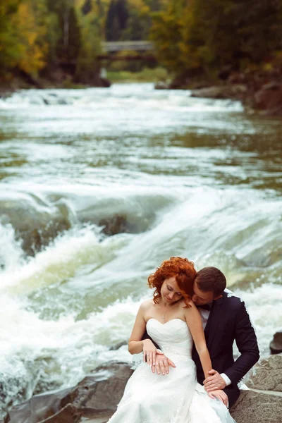 Mariée et marié assis sur les pierres tandis qu'une rivière de montagne coule b — Photo