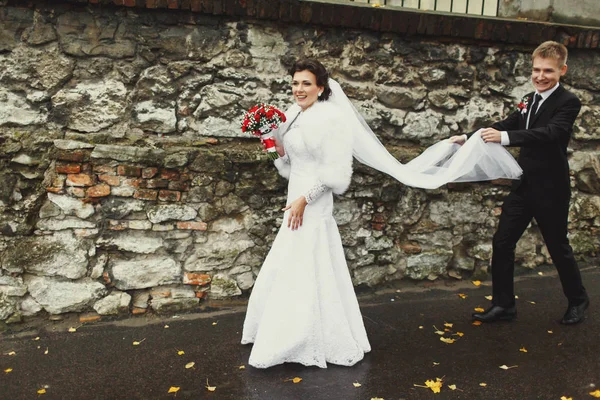 Groom segura o véu da noiva enquanto caminham ao longo da rua molhada — Fotografia de Stock