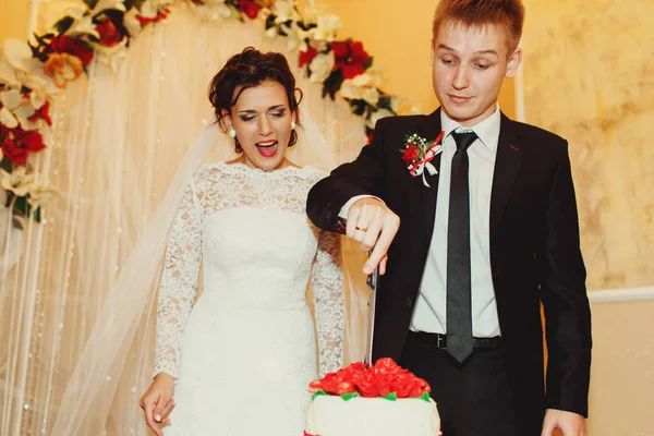 Жених режет свадебный торт, а невеста смеется позади него — стоковое фото