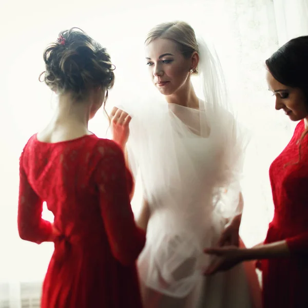 As damas de honra bonitas em vestidos vermelhos ajustam um véu em uma noiva — Fotografia de Stock