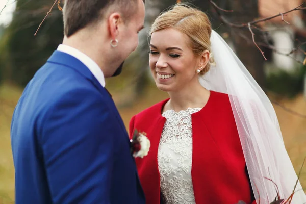 Groom fica atrás de uma noiva sorridente incrível em uma jaqueta vermelha — Fotografia de Stock