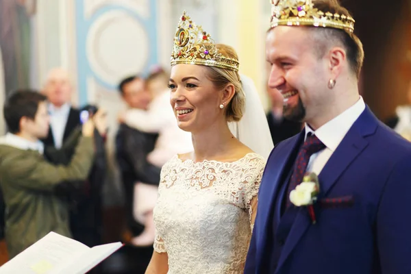 Noiva e noivo rir enquanto está em coroas atrás de um padre — Fotografia de Stock