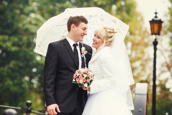 Braut und Bräutigam lächeln umarmt unter einem weißen Regenschirm im Regen — Stockfoto