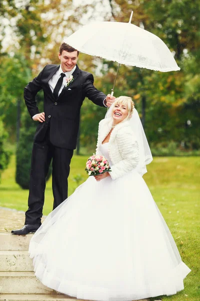 Mariée et marié s'amusent à marcher avec un parapluie autour du pair — Photo