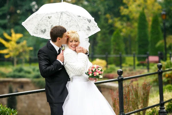 Sposo bacia la testa della sposa mentre lei si appoggia a lui sotto un ombrello — Foto Stock