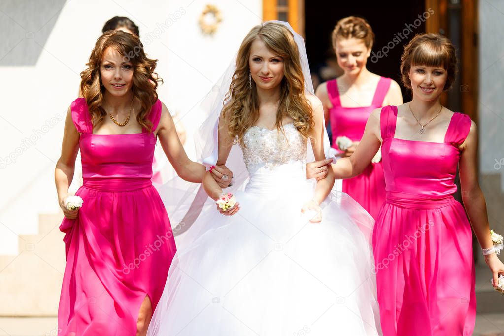 Bridesmaids in pink dresses hold bride's arms walking out of the