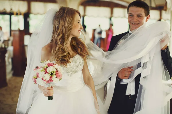 Groom holds bride's veil walking with her in an empty restaurant — Stock Photo, Image