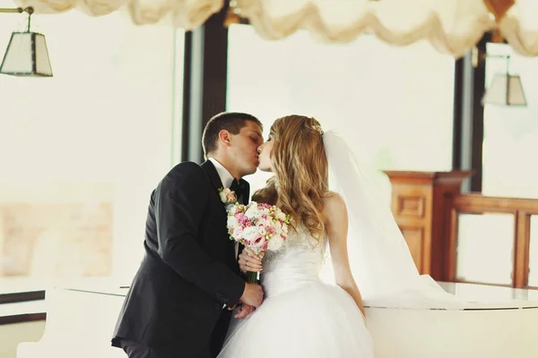 Os recém-casados beijam-se apoiando em um piano — Fotografia de Stock