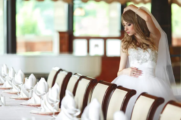 Delgada novia ajusta su cabello rubio posando detrás de una mesa vacía — Foto de Stock