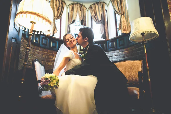 Groom leans bride to himself kissing her cheek in a vintage hall — Stock Photo, Image