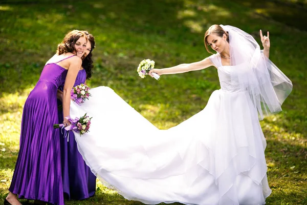 Bride makes rock'n'roll while pretty bridesmaids carry her dress — Stock Photo, Image