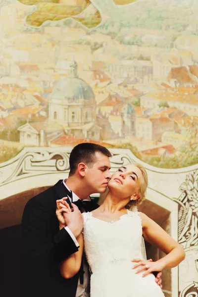 Recién casados beso de pie detrás de una pared con ciudad al aire libre — Foto de Stock