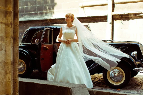 Ветер развевает завесу невесты, пока она шагает по ретро-машине — стоковое фото