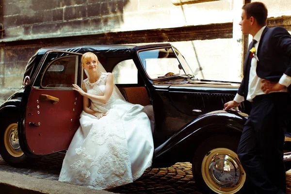 Bride sits in an open door of a retro car while groom waits behi — Stock Photo, Image