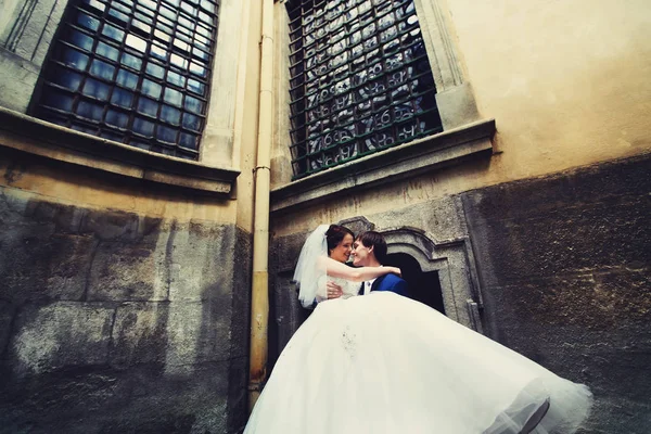 Novio sostiene novia en sus brazos de pie bajo grandes ventanas — Foto de Stock