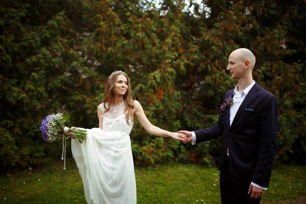 Novia sostiene un ramo y un vestido en un brazo y la mano del novio en — Foto de Stock