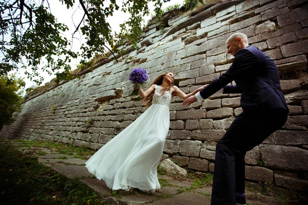 Bruid wervelingen aan een bruidegom langs een pad achter een oude stenen muur — Stockfoto