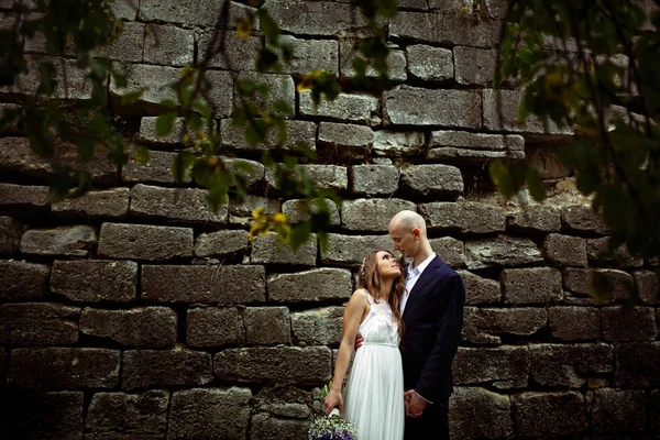 Bride looks in groom's eyes while they stand behind an old stone — Stock Photo, Image