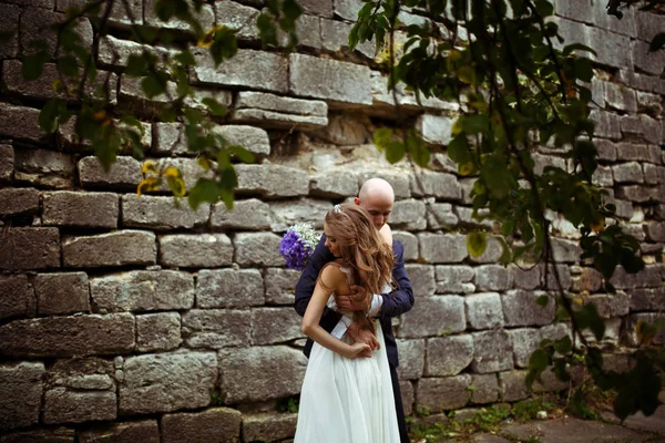 Vento sopra o cabelo da noiva enquanto o noivo a abraça debaixo da árvore — Fotografia de Stock