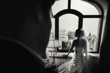 Büyük bir pencere önünde duran bir damat gelin bekler 