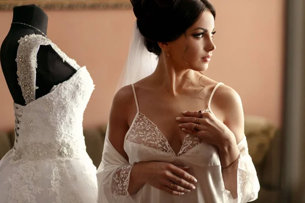 La novia encantadora se encuentra cerca del vestido de novia en la habitación —  Fotos de Stock