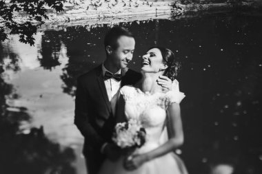 Damadın hugs bir bahçe göl arkasında duran gelin gülümsüyor 