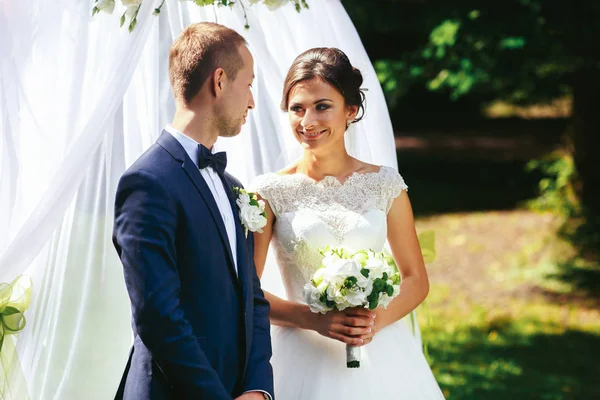 Los recién casados se ven divertidos de pie en la parte delantera del altar blanco en el — Foto de Stock