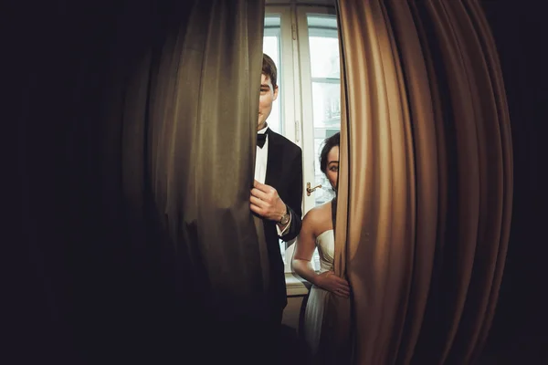 Bride and groom hide behind the curtains in a hotel room — Stock Photo, Image