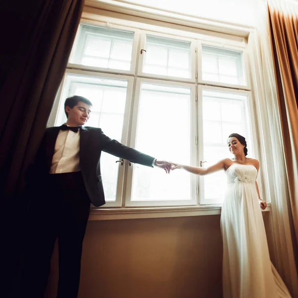 Groom segura a mão da noiva de pé com ela atrás de uma janela — Fotografia de Stock