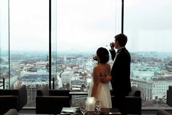 Los recién casados beben champán mirando por la ventana a gran ci — Foto de Stock