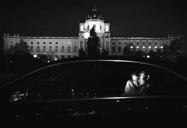 Noiva se inclina para um noivo sentado no carro com uma grande construção velha — Fotografia de Stock
