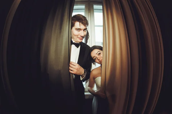 Funny newlyweds stand behind the beig curtains — Stock Photo, Image