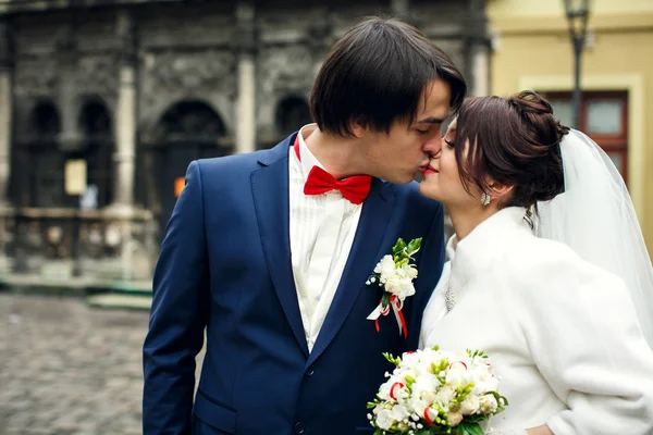 Groom kisses a brunette bride standing with her on a city square — Stock Photo, Image