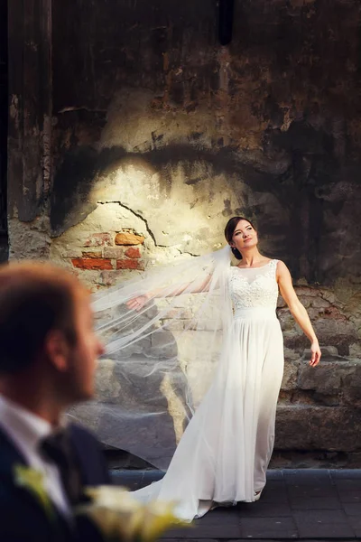 Groom admira uma noiva posando atrás de uma parede de pedra velha — Fotografia de Stock