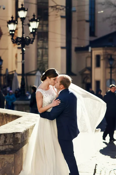 El viento sopla un velo alrededor de los recién casados mientras se besan en la vieja plaza — Foto de Stock