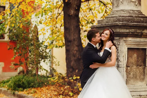 Recém-casados sorriem atrás de um velho pilar cinzento — Fotografia de Stock