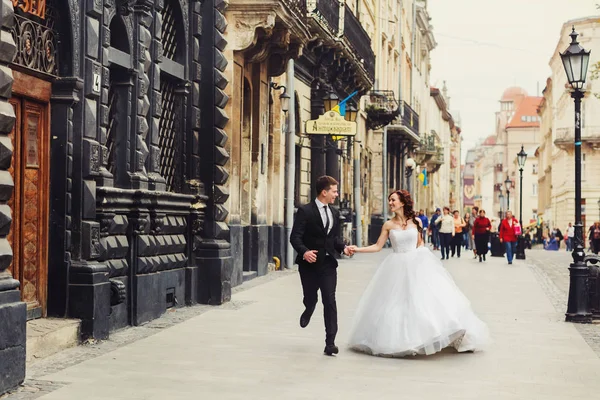 Casal de casamento feliz corre ao longo da rua velha com grande arco — Fotografia de Stock