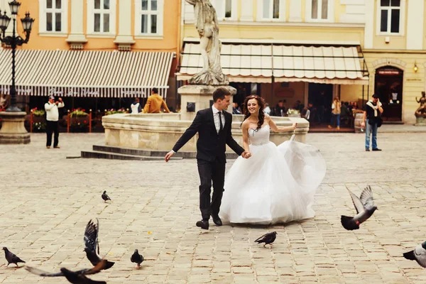 Groom segura a mão da noiva enquanto eles correm através de uma cidade quadrada ti — Fotografia de Stock