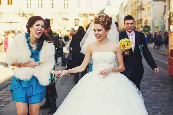Pretty bride and bridesmaid wlak along the street holding their — Stock Photo, Image