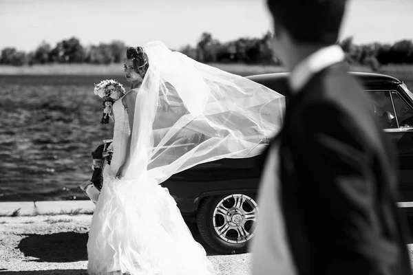 El viento sopla velo de novia mientras ella está detrás de un coche viejo — Foto de Stock