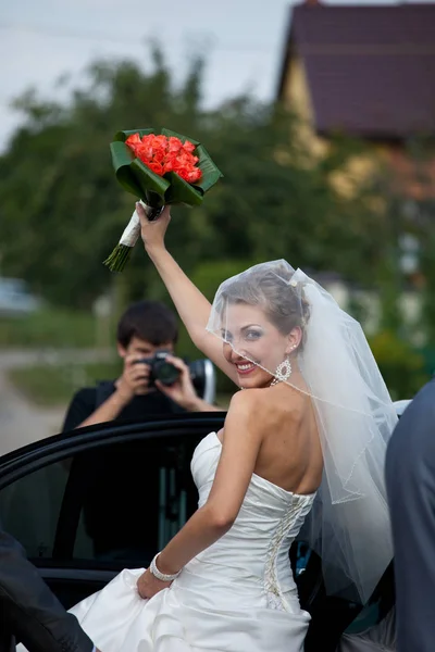 Smiling bride raises a hand with a bouquet sitting in the car — Stock Photo, Image