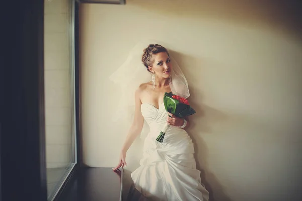 Gorgeous bride poses with red bouquet behind a window — Stock Photo, Image