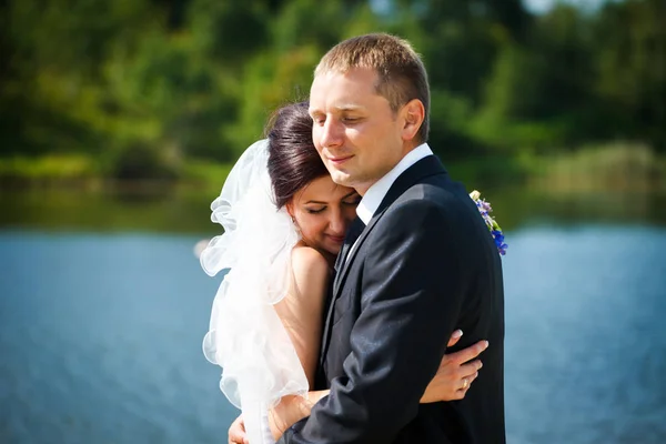 Braut lehnt sich eng an einen Bräutigam, der vor einem See steht — Stockfoto