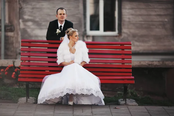 Ženich stojí za červeném lavičce, zatímco nevěsta sedí na něm — Stock fotografie