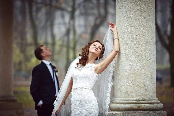 Mooie bruid met krullend haar neigt naar een marmeren pijler terwijl g — Stockfoto