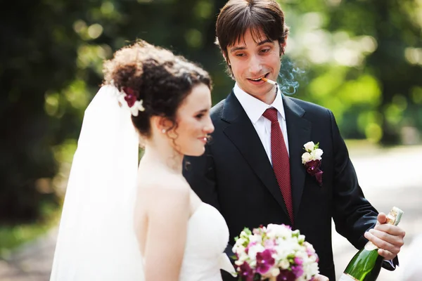 Rauchender Bräutigam steht hinter einer Braut mit einer Flasche Champagner — Stockfoto